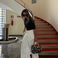 Hanna MW wearing a black t-shirt, white trousers, and a Louis Vuitton Speedy bag next to a red spiral staircase.