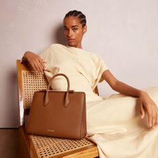 a model poses with a brown top-handle bag by Strathberry