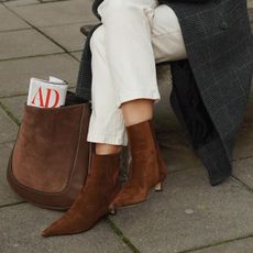 Woman sitting on bench wearing brown suede boots with grey coat and white jeans.