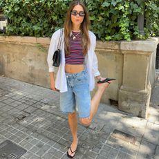 Dutch fashion influencer Luna Isabella on the street during summer wearing an oversize white shirt open over a cropped striped tank, long denim shorts, kitten-heeled black flip-flops, rectangular sunglasses, a Chanel shoulder bag, and a gold heart necklace.